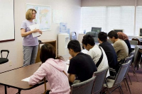 Photograph of a classroom setting with a teacher standing and holding papers while five students sit at a table facing the teacher. 