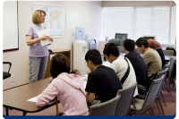 Photograph of a classroom setting with a teacher standing and holding papers while five students sit at a table facing the teacher. 