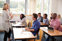 Photograph of a classroom setting with a teacher standing and speaking to a group of adult students seated at desks