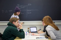 Photograph of four students engaged in a group study session around a table with a laptop, notebooks, and pens. 