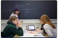 Photograph of four students engaged in a group study session around a table with a laptop, notebooks, and pens. 