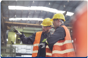 Photograph of two construction workers wearing yellow hard hats and orange reflective vests inside an industrial facility